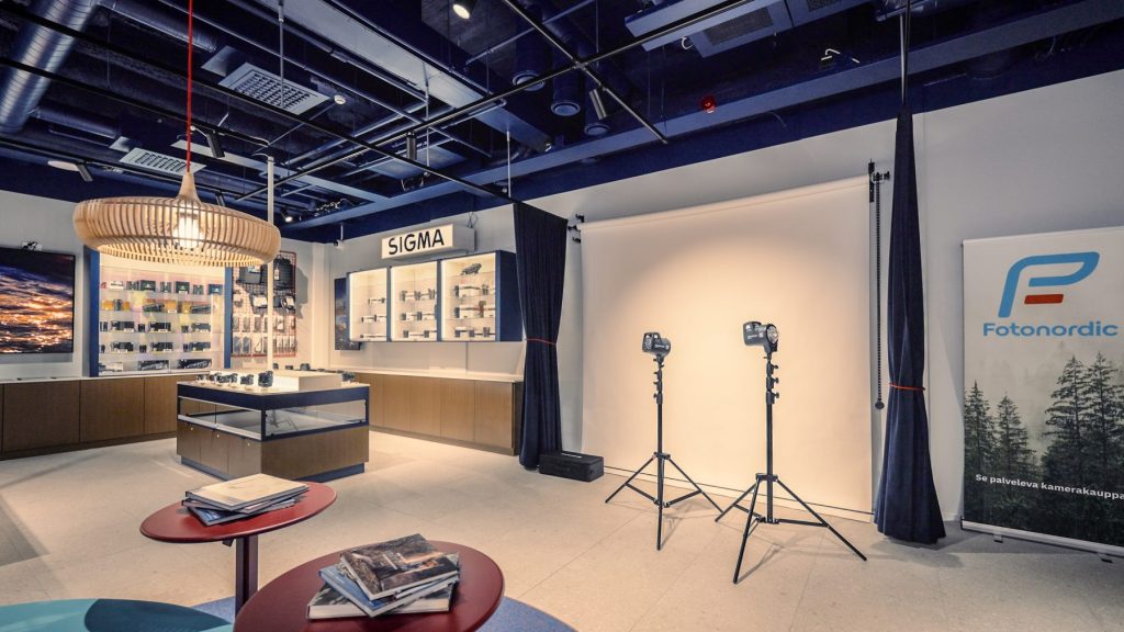 Fotonordic.fi liiketila Helsinki mikonkatu 7 Sisu interior sisustusarkkitehtitoimisto. Tummansiniseksi maalattu alakatto, jossa näkyy talotekniikka. Valokuvausstudio, jossa valkoinen backdrop kangas telineessä, jalalliset studio valot sekä siniset pitkät verhot. Tuotemerkki Sigman kameroita.